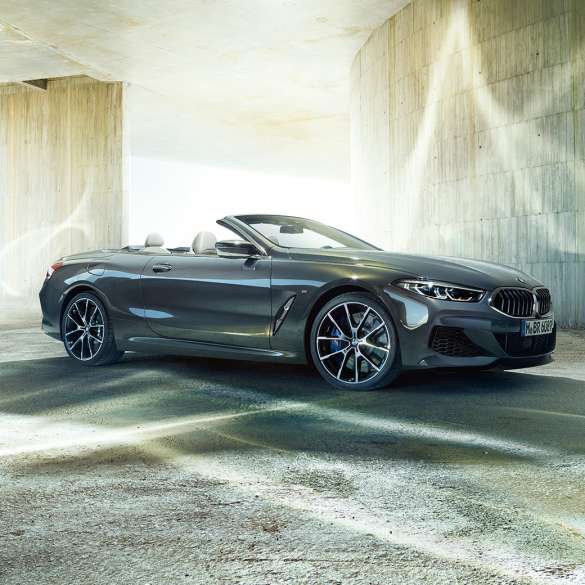 BMW M850i xDrive Convertible, three-quarter side view. BMW M850i xDrive Convertible in Dravit Grey metallic with open softtop, three-quarter side view, standing in front of concrete building.