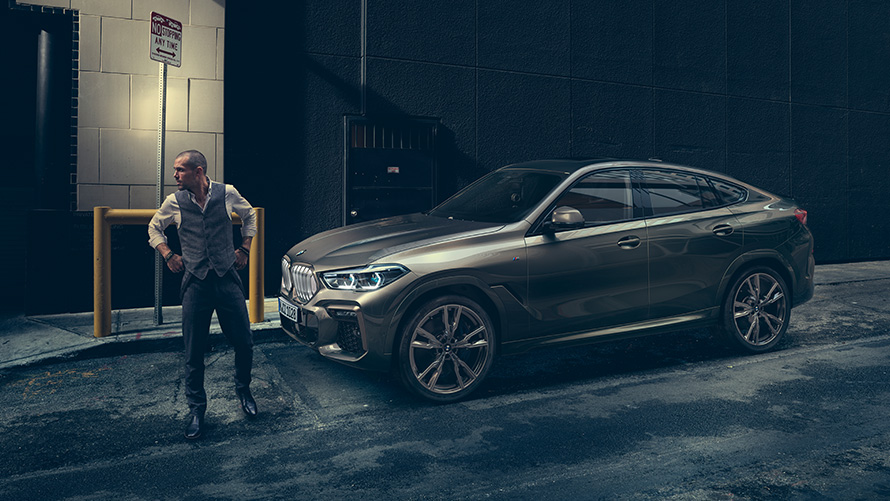 Man standing in front of BMW X6 M50i in lateral perspective in front of an urban setting.