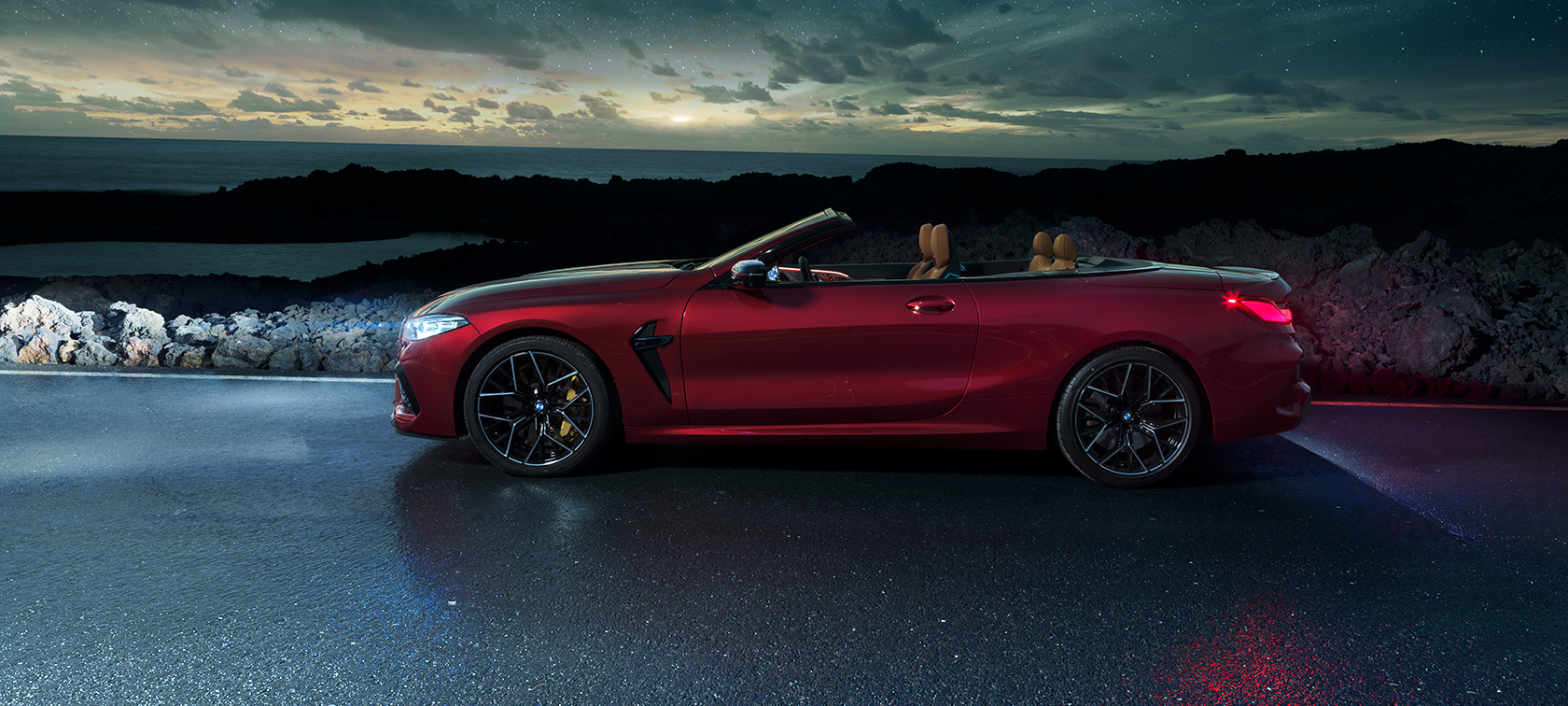 BMW M8 Competition Convertible, side view. BMW M8 Competition Convertible in Motegi Red metallic with open softtop, side view, standing on coast road under starry sky.