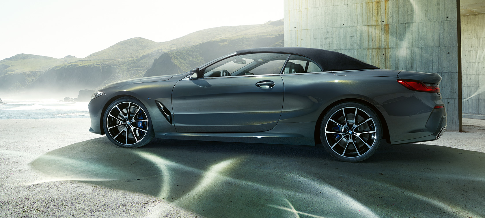 BMW M850i xDrive Convertible, side view. BMW M850i xDrive Convertible in Dravit Grey metallic with closed softtop, side view in front of hills.