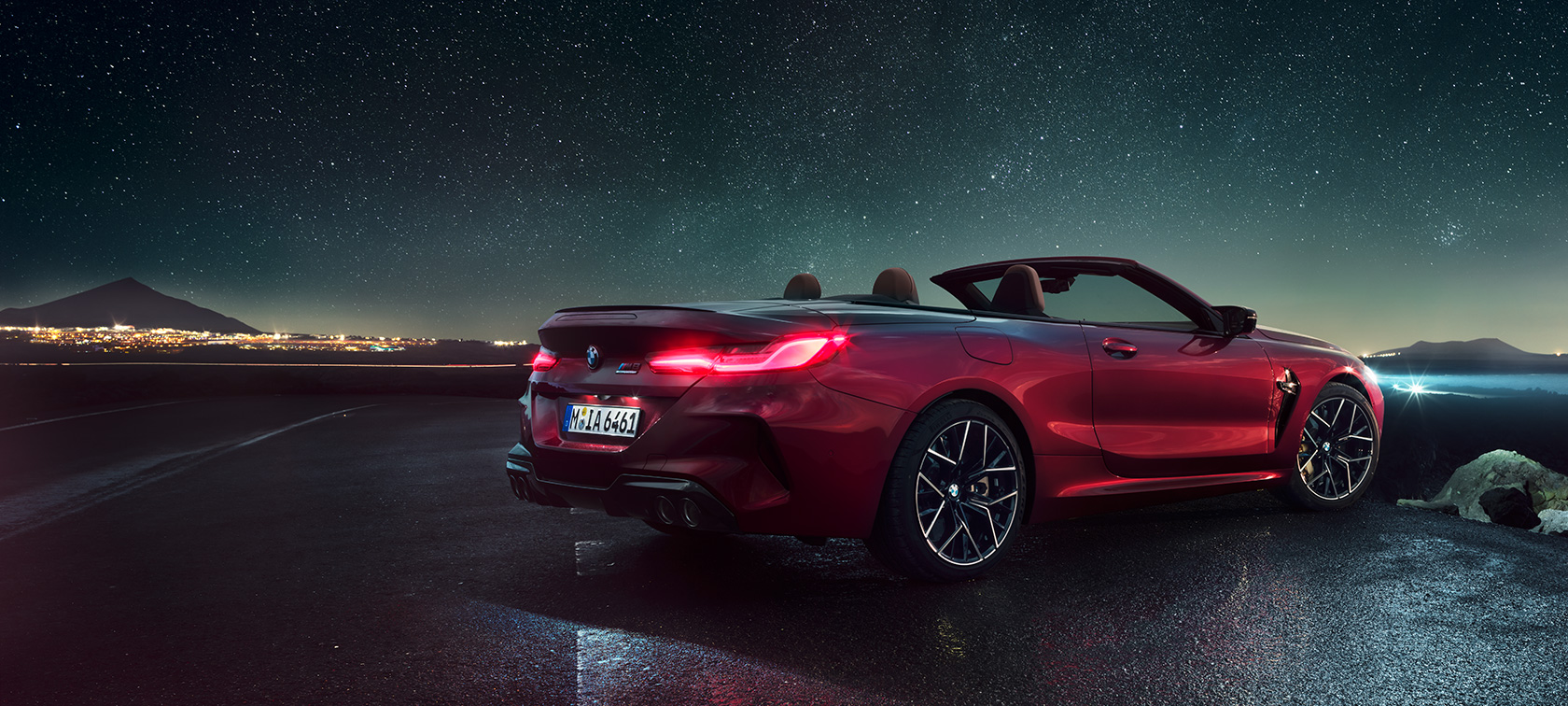 BMW M8 Competition Convertible, three-quarter rear view. BMW M8 Competition Convertible in Motegi Red metallic with open softtop, three-quarter rear view, standing under starry sky.
