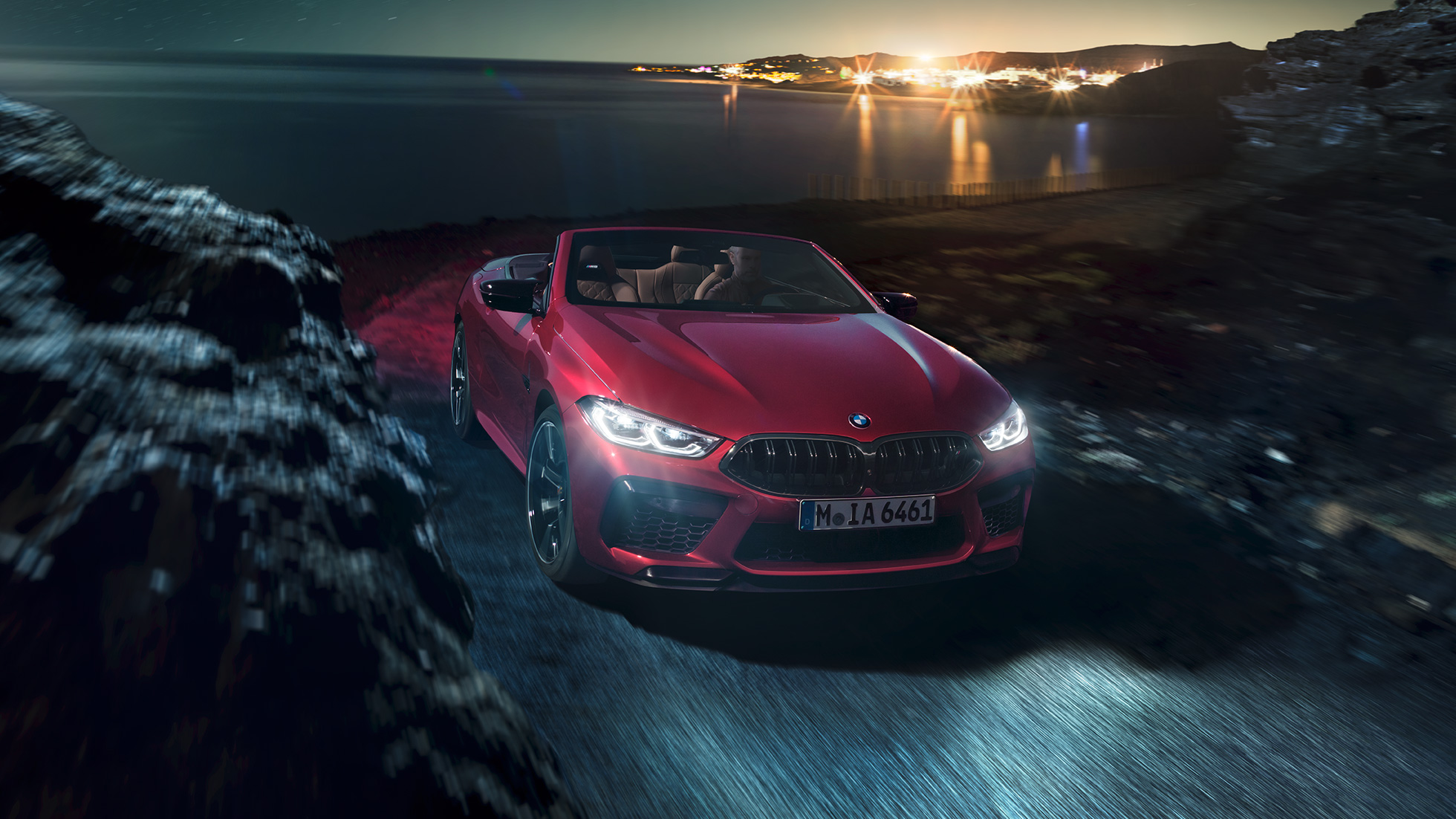 BMW M8 Competition Convertible, three-quarter front view. BMW M8 Competition Convertible in Motegi Red metallic with open softtop, three-quarter front view, driving at night on coast road.