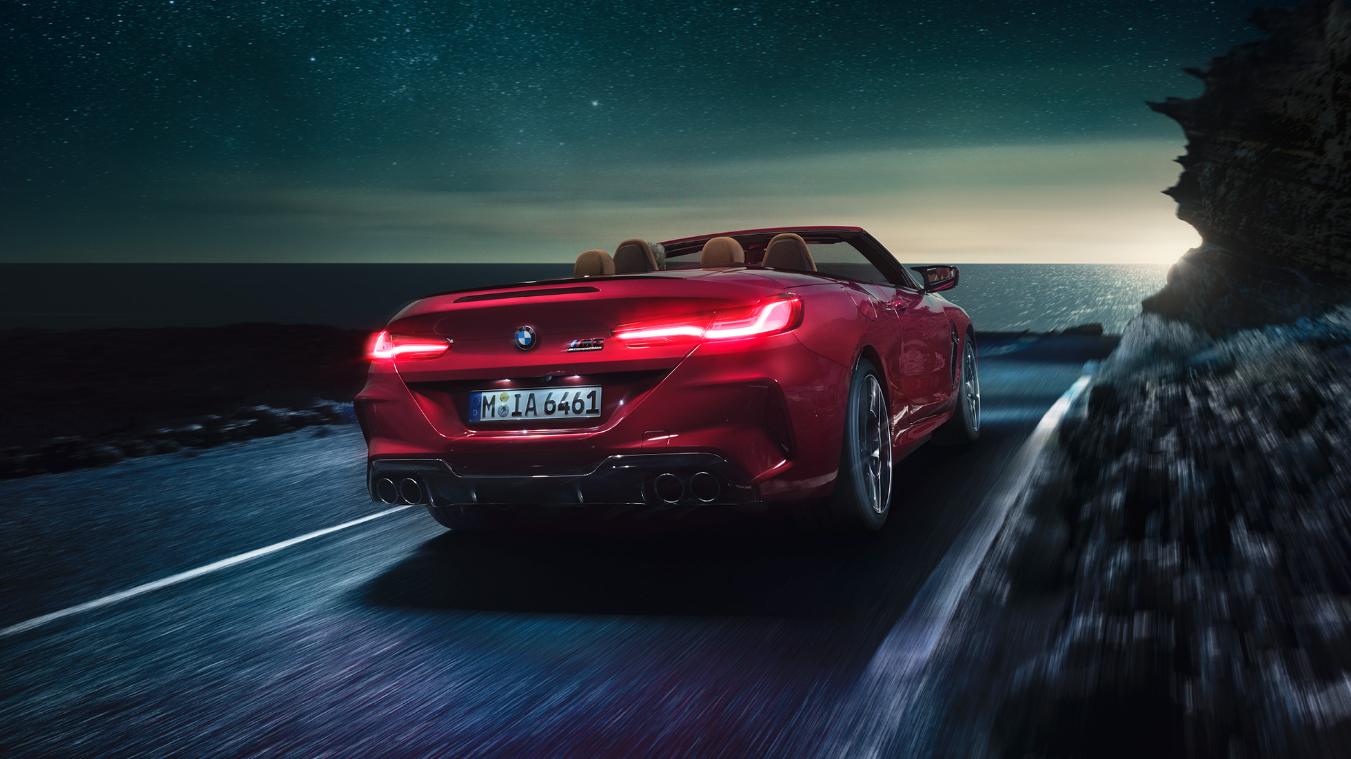 BMW M8 Competition Convertible, three-quarter rear view. BMW M8 Competition Convertible in Motegi Red metallic with open softtop, three-quarter rear view, driving at night on coast road.