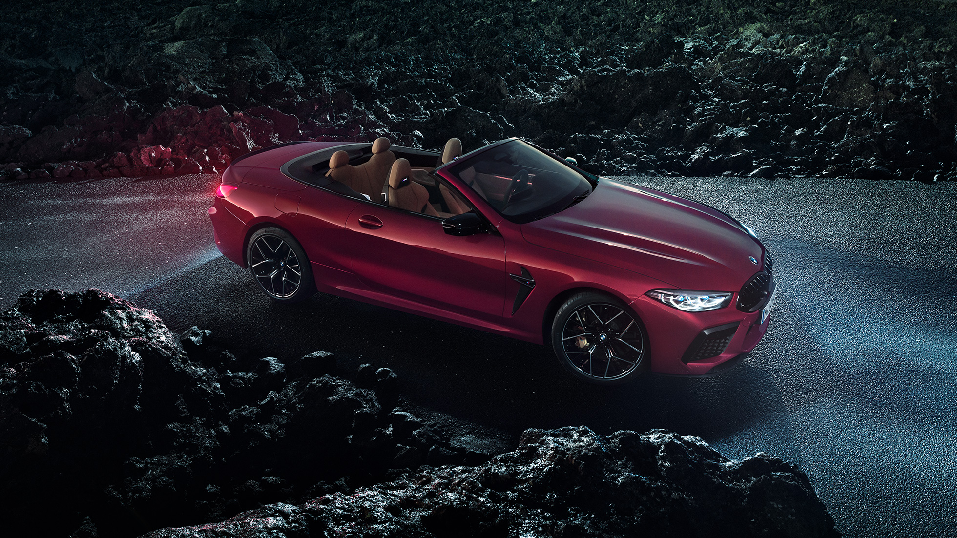 BMW M8 Competition Convertible, three-quarter side view. BMW M8 Competition Convertible in Motegi Red metallic with open softtop, three-quarter side view, standing on asphalt at night.