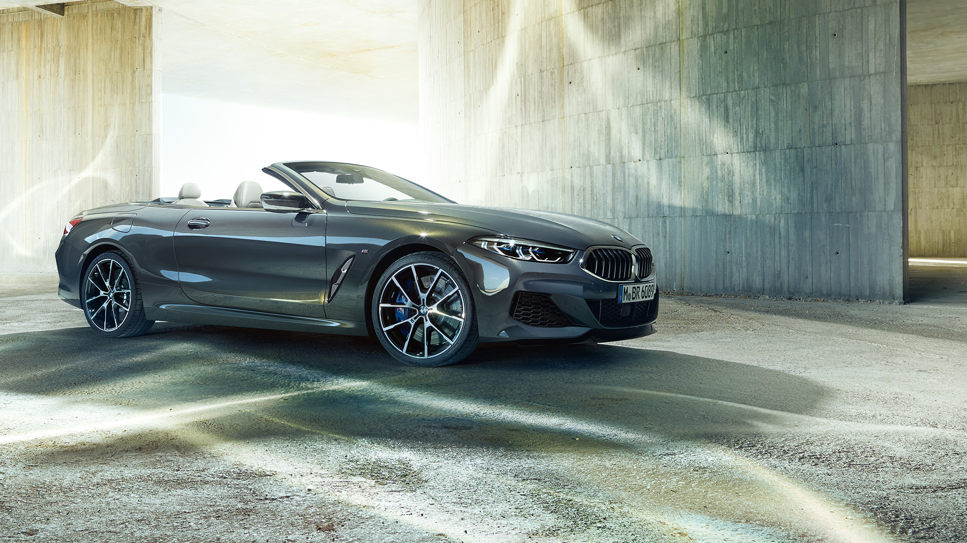 BMW M850i xDrive Convertible, three-quarter side view. BMW M850i xDrive Convertible in Dravit Grey metallic with open softtop, three-quarter side view, standing in front of concrete building.