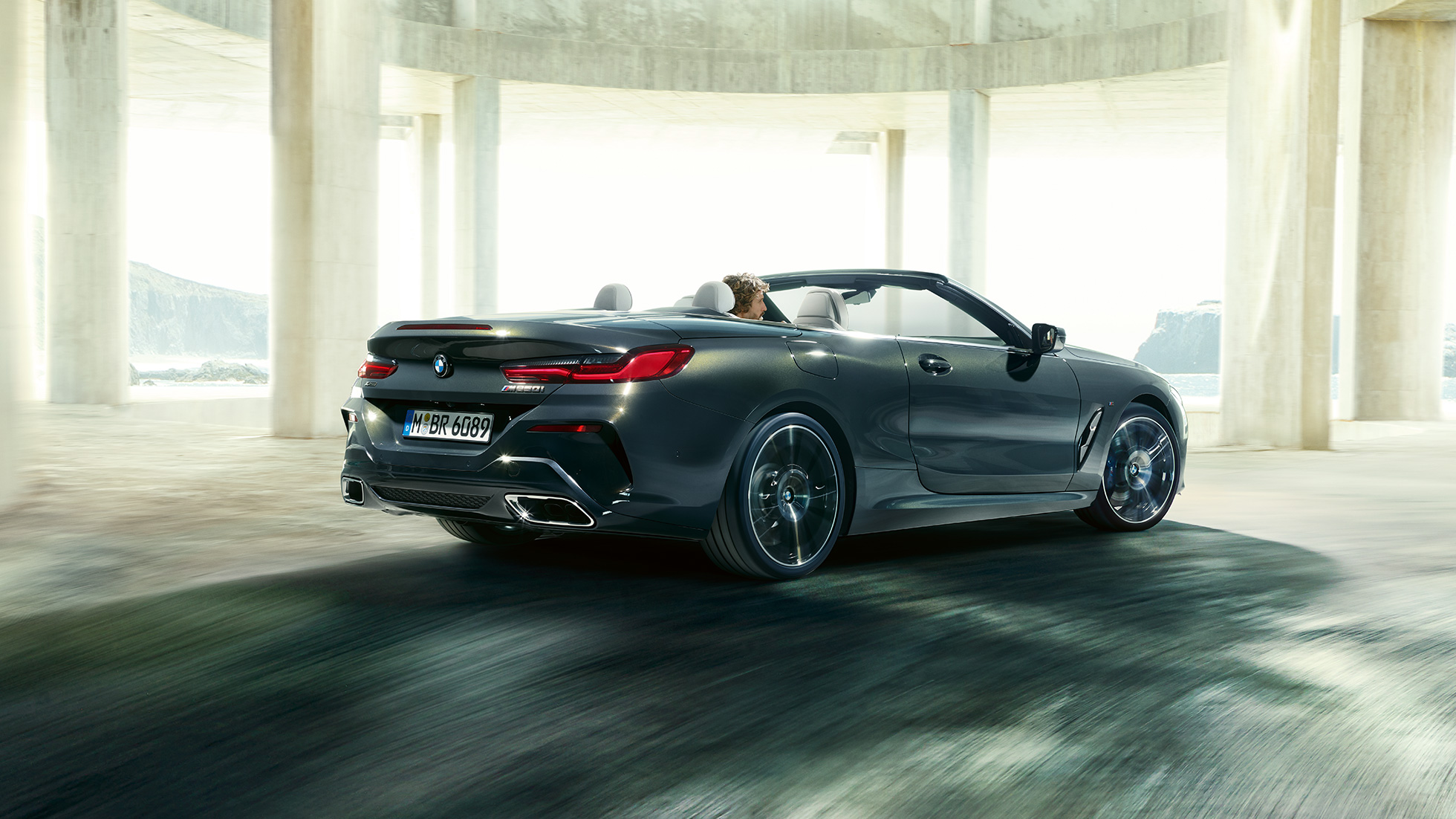 BMW M850i xDrive Convertible, three-quarter rear view. BMW M850i xDrive Convertible in Dravit Grey metallic with open softtop, three-quarter rear view, driving through concrete building.