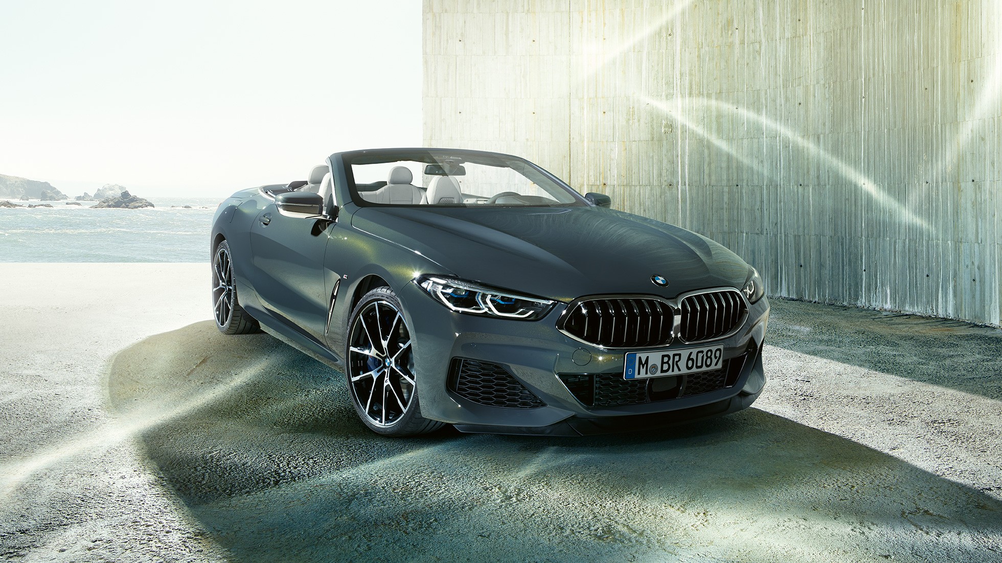 BMW M850i xDrive Convertible, three-quarter front view. BMW M850i xDrive Convertible in Dravit Grey metallic with open softtop, three-quarter front view, standing in front of coast.