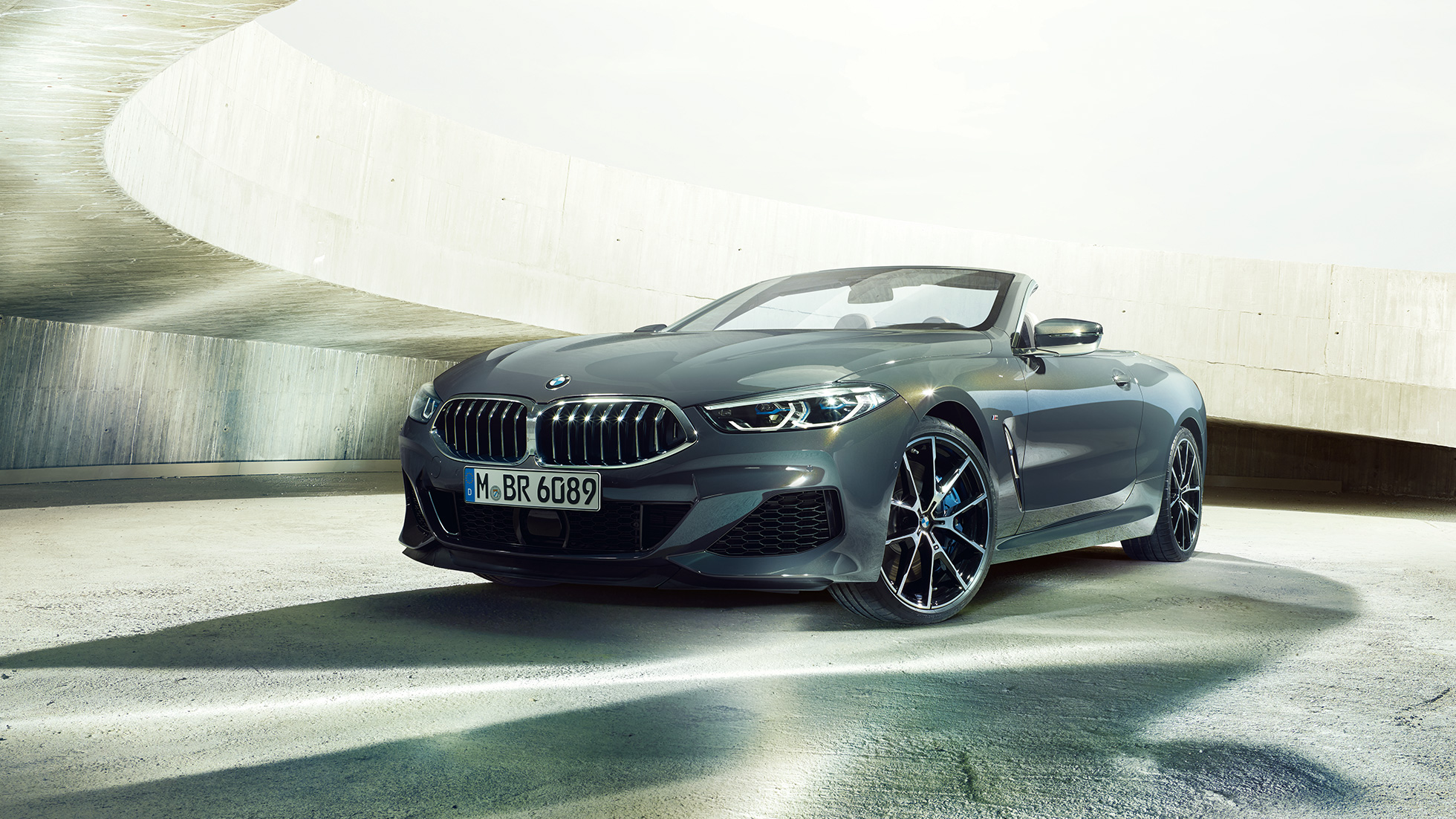 BMW M850i xDrive Convertible, three-quarter front view. BMW M850i xDrive Convertible in Dravit Grey metallic with open softtop, three-quarter front view, standing in concrete building.