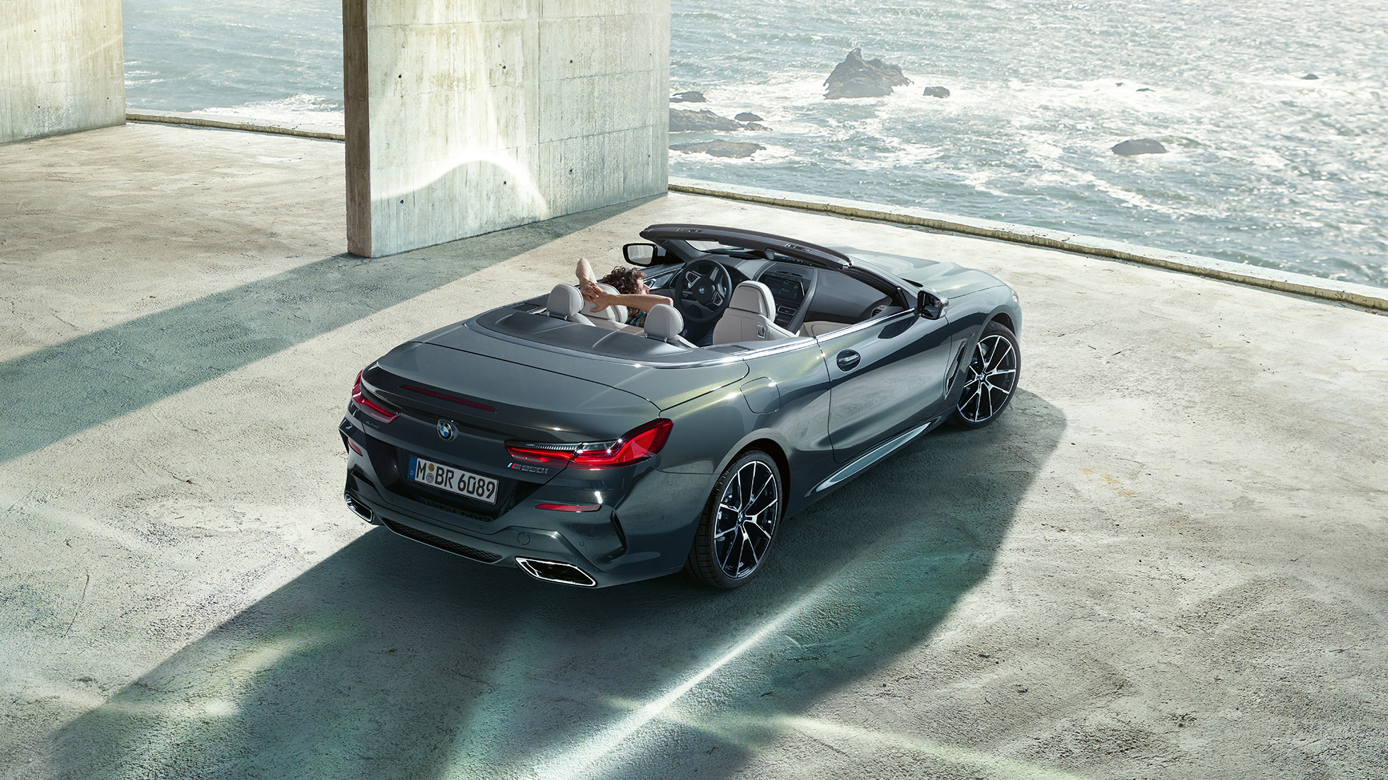 BMW M850i xDrive Convertible, bird's eye view. BMW M850i xDrive Convertible in Dravit Grey metallic with open softtop, bird's eye view, parking in concrete building in front of ocean.