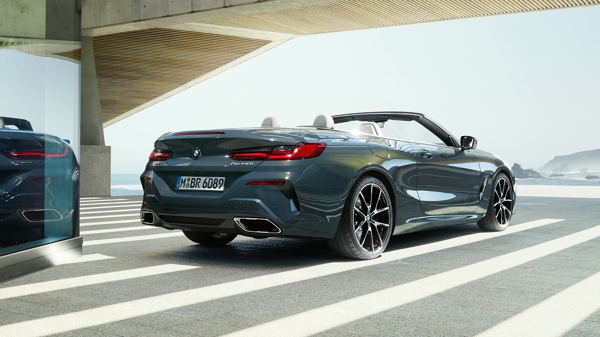 BMW M850i xDrive Convertible, three-quarter rear view. BMW M850i xDrive Convertible in Dravit Grey metallic with open softtop, three-quarter rear view, parking with ocean view.
