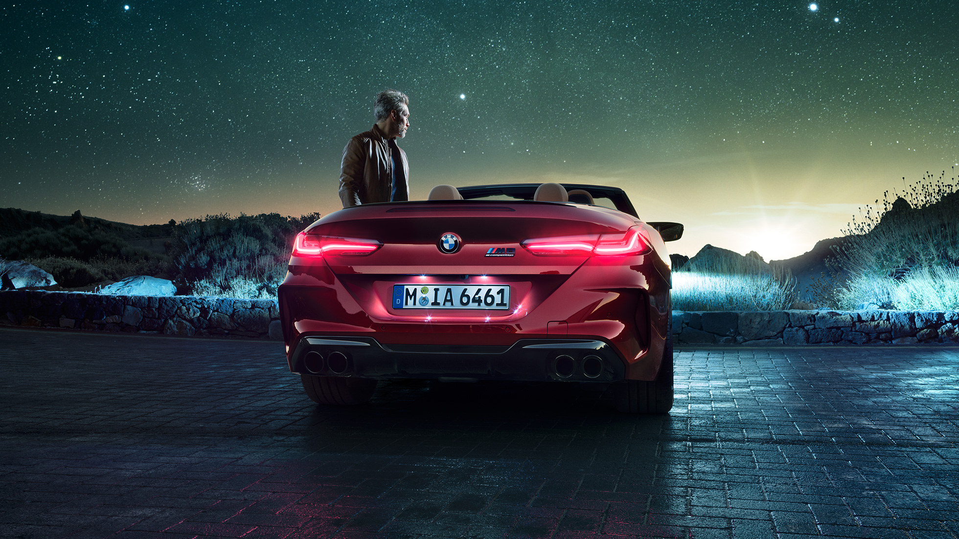 BMW M8 Competition Convertible, rear view. BMW M8 Competition Convertible in Motegi Red metallic with open softtop, rear view, standing with man under starry sky.