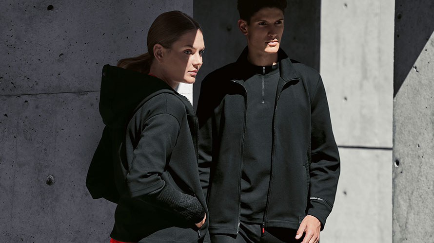 A couple ist standing in the shadow. A woman is presenting the BMW M Sweat Jacket for ladies and a man is wearing the BMW M Sweat Jacket for men.