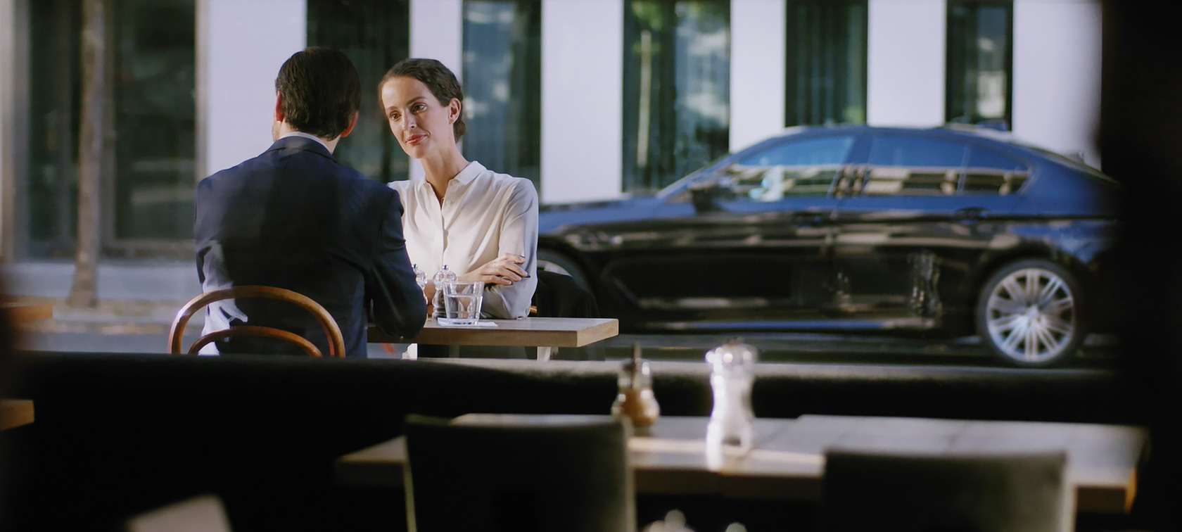 BMW Corporate Aftersales, man and woman sitting in a café BMW Corporate Aftersales: Man and woman sitting in a café, a BMW in the background